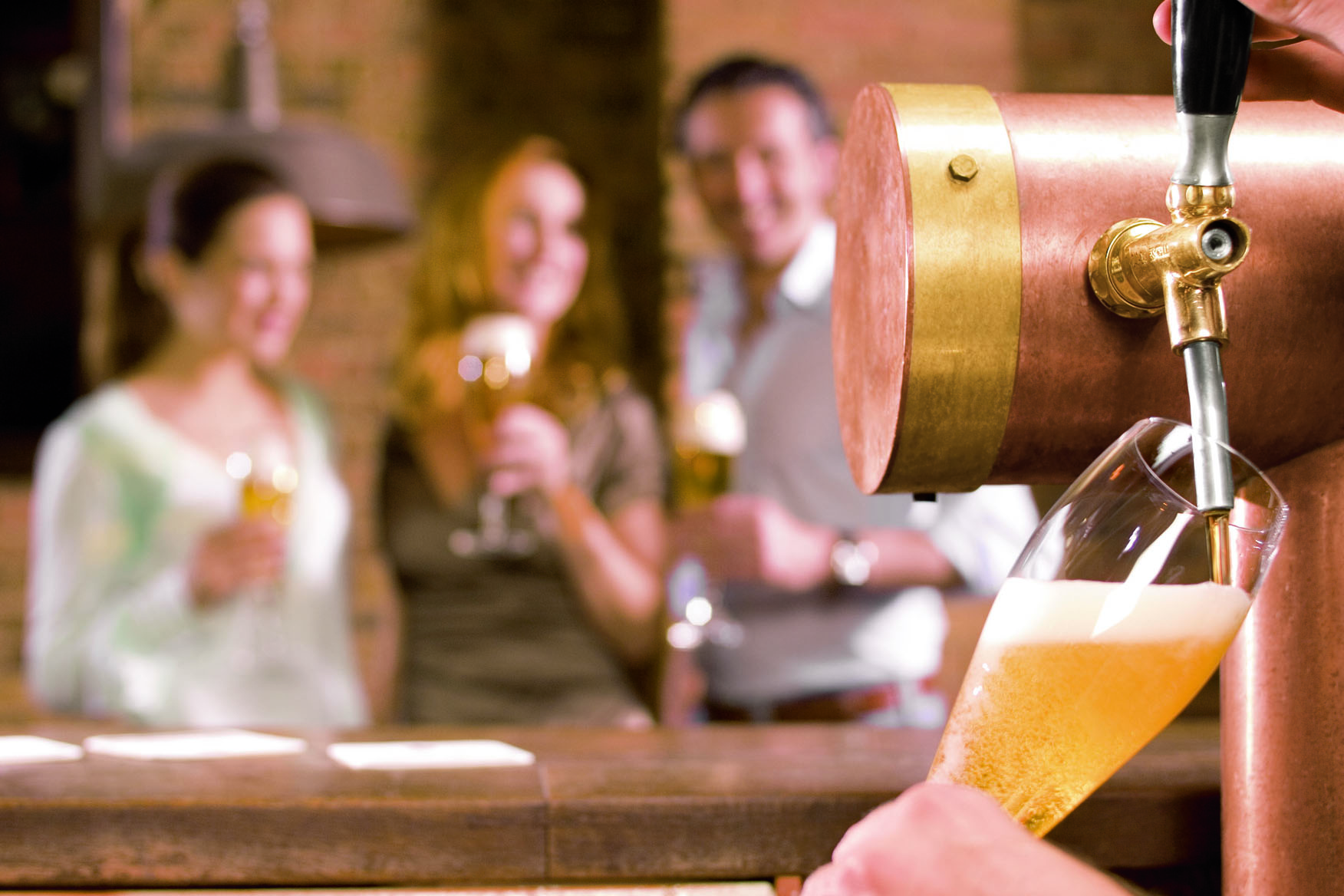 Ein Bier wird frisch vom Fass in ein Glas gezapft, im Hintergrund lachen Menschen.